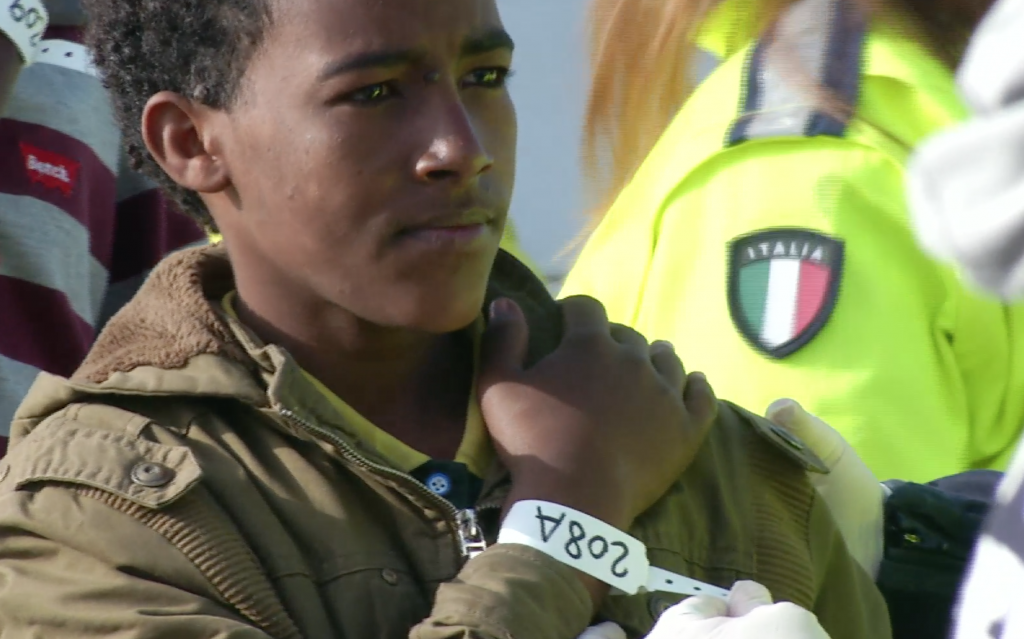 A young migrant with a number, 208A, looks determined and hopeful as he stands in the port of Pozzallo, Sicily after being rescued at sea. Freeze frame of video shot by AP Television Cameraman Luigi Navarra April 17, 2015